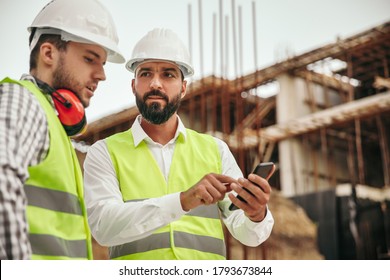 Low angle of professional male technicians in workwear and hardhats using smartphone while discussing engineering plan on construction site of contemporary building