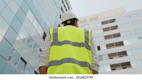 Low Angle Rear View Of Engineer Looking Up At Modern Building Outdoors. Male Architect In Safety Vest And Helmet Standing Outside At Construction Site