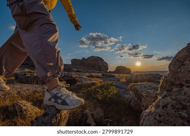 Low angle rear view of solo traveler walking up rocky mountain hill at sunset. Close up of sneaker boot exploring remote wilderness steppe landscape during golden hour.