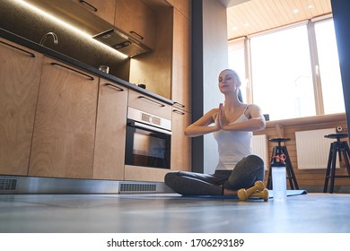 Low Angle Of A Serene Young Female Yogi Sitting In A Prayer Pose On The Kitchen Floor