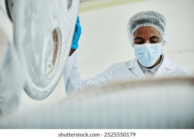 Low angle shot of African American male technician in protective mask looking inside steel tank opening large lid while inspecting production process at pharmaceutical manufacturing plant, copy space - Powered by Shutterstock