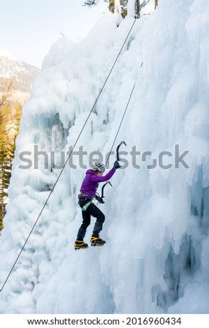 Low angle shot of an alpinist woman with ice climbing equipment, axe and climbing ropes, climbing at a frozen waterfall, view from below