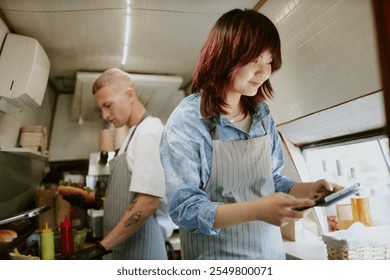 Low angle shot of Asian female cook checking new orders in app on tablet while Caucasian male chef preparing hot dogs in blurred background - Powered by Shutterstock