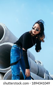 Low angle shot of black hair girl in sweater and jeans standing and bend down her body on the big concrete tube.