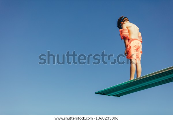 Low angle shot of boy with sleeves floats on diving board preparing for dive in the pool. Boy standing on diving spring board against sky.