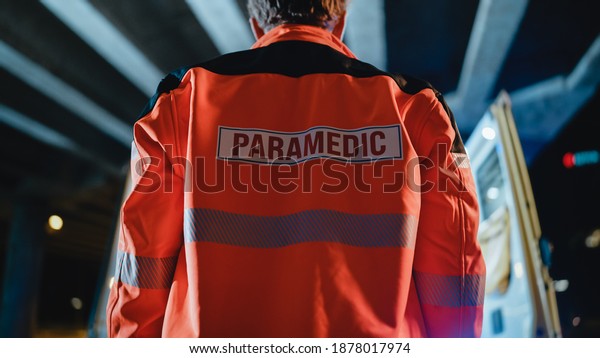 Low Angle Shot of a EMS Paramedic Proudly\
Standing With His Back Turned to Camera in High Visibility Medical\
Orange Uniform with Paramedic Text Logo. Successful Emergency\
Medical Technician or\
Doctor.