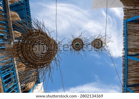A low angle shot of ethnic rustic lamps made from woven palm fibers or straw hanging between traditional buildings with blue wooden railings.
