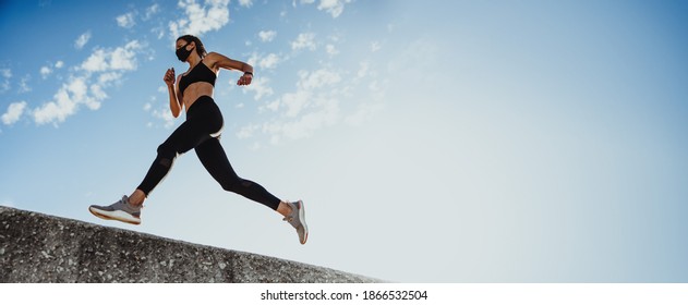 Low angle shot of a female athlete doing running workout over a wall against blue sky. Panoramic shot of a female in sportswear and protective face mask exercising in morning. - Shutterstock ID 1866532504