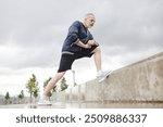 Low angle shot of fit elderly man in sportswear stretching leg muscles in lunge position on concrete steps against cloudy sky in city park, copy space