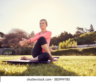 Low angle shot of a fit senior woman outdoors on a summer morning, with her yoga mat on the grass, doing a twist exercise to stretch her back