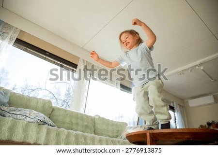 Low angle shot of joyful small child with long hair jumping and soaring off sofa, full of energy and playful laughter during a bright morning at home