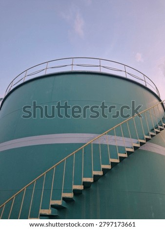 ​A low angle shot of a large cylindrical turquoise metal storage tank or industrial silo featuring yellow external spiral stairs against a soft twilight sky.