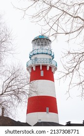 A low angle shot of a lighthouse surrounded by leafless branches of trees against a clear white cloudy sky. Early spring or winter