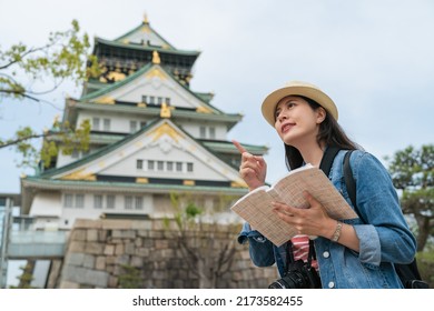 Low Angle Shot Portrait Of Cheerful Asian Female Tourist Finger Pointing Into Distance While Exploring In Osaka Castle Park In Japan With Travel Guidebook In Hand