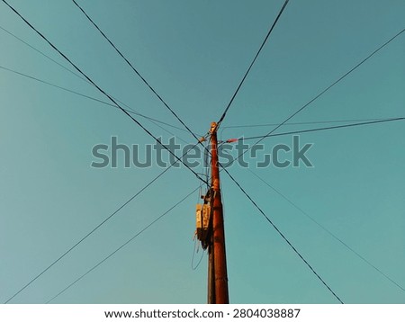 Low angle shot of a single utility pole with power lines and internet cables spreading out in a cross pattern against a clear cyan sky. Minimalist composition featuring a distribution junction box on a concrete pylon during golden hour.