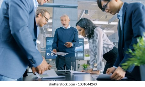 Low Angle Shot Of The Standing Group Of Successful Business People In The Conference Room, Work On A Company's Growth, Share Charts And Statistics. In The Background Wall TV With Corporate Data On It.