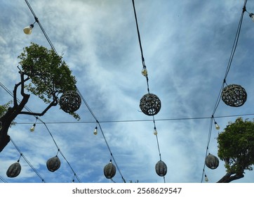 A low angle shot of a string of woven light bulbs hanging against a blue sky with white clouds. Several trees are visible on either side of the image.