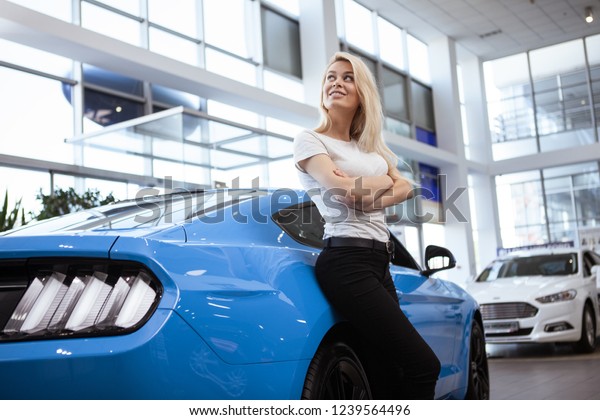 Low angle shot of stunning young female driver\
posing with her new automobile at the dealership salon. Happy woman\
looking away dreamily, leaning on a modern muscle car, copy space.\
Buying car concept