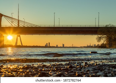 Low Angle Shot Towards City Riga During Sunset. In Front River Daugava Has Beautiful Golden Bokeah Of Sun Shining Straight To It. Back Of The Photo Is South Bridge, TV Tower And Panorama Of The Riga.