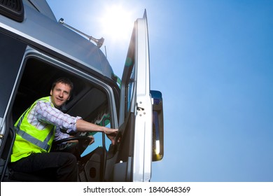 A Low Angle Shot Of A Truck Driver In Safety Vest Sitting Behind The Wheel Inside The Cab Of A Semi-truck On A Bright Sunny Day While Closing The Door.