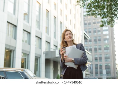 Low angle shot of young elegant caucasian businesswoman in formal wear holding takeaway coffee cup walking outside office during the break with laptop and documents. Office worker at business center. - Powered by Shutterstock