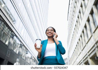 Low Angle Shot Of Young Elegant African American Business Woman In Formal Wear Talking On Phone And Holding Takeaway Coffee Cup While Walking Outside Office During The Break