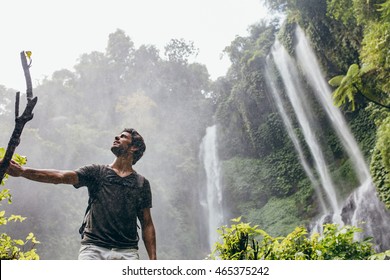 Low angle shot of young man standing near a waterfall in forest and looking away. Male hiker enjoying in the nature with rain.