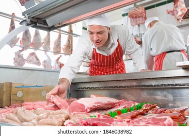 A low angle shot of a young smiling butcher in a red apron arranging meat in a display case. - Powered by Shutterstock