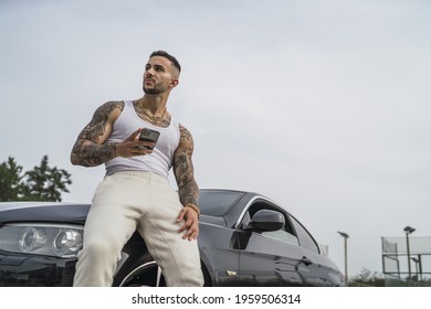 A Low Angle Shot Of A Young Tattooed Man Leaning On His Car And Holding His Phone