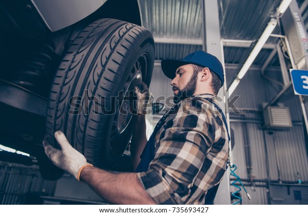 Low angle side\
profile shot of expert specialist technician changes tires, tyres\
of lifted up car, at auto workshop, wears checkered shirt, uniform\
costume, hat headwear
