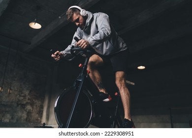 Low angle side view of bearded male exercising on stationary cycle in dark gym while holding handle bar tightly and looking down while stepping on pedals with legs - Powered by Shutterstock