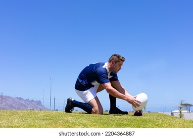 Low angle side view of a Caucasian male rugby player wearing a team uniform, training at a sports field, kneeling and placing a rugby ball for a kick, with blue sky in the background - Powered by Shutterstock