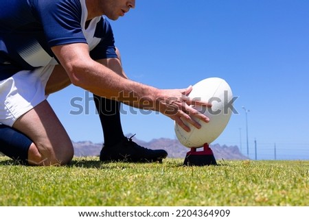 Low angle side view close up of a Caucasian male rugby player wearing a team uniform, training at a sports field, kneeling and placing a rugby ball for a kick, with blue sky in the background
