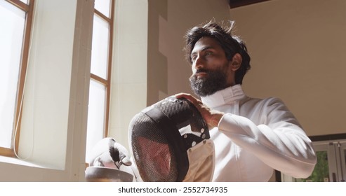 Low angle side view of a confident focused biracial male fencer athlete during a fencing training in a gym, wearing jacket and plastron, preparing for a fencing duel, putting mask on, holding an epee