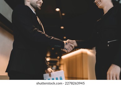 Low angle side view of crop anonymous smiling male and female colleagues in black suits standing in modern office hallway and shaking hands after meeting
