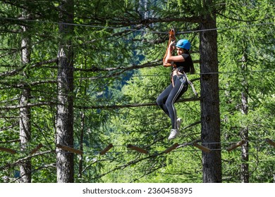 Low angle side view of excited young female in helmet while hanging from top rope with safety belay cables and walking on suspended wire in forest adventure park in daylight - Powered by Shutterstock