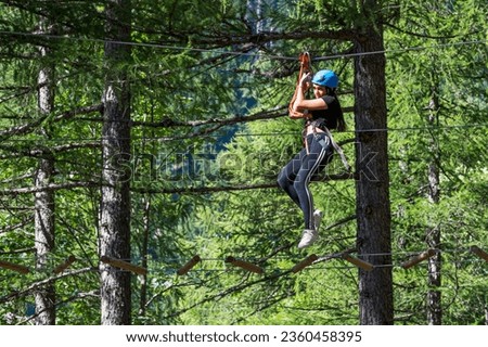Low angle side view of excited young female in helmet while hanging from top rope with safety belay cables and walking on suspended wire in forest adventure park in daylight