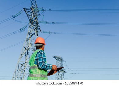 Low angle side view of focused young electrical engineer in hardhat standing near modern high voltage power line and examining papers