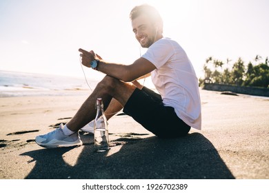 Low angle side view of glad male in sportswear listening to music in earphones while sitting with bottle of water on sandy seashore and looking at camera in back lit