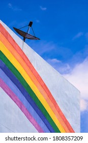 Low angle and side view of satellite dish on top of white building with rainbow stripes on building wall against blue sky background 