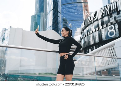 Low angle side view of slim female athlete in sportswear taking self portrait on cellphone while listening to music against modern buildings