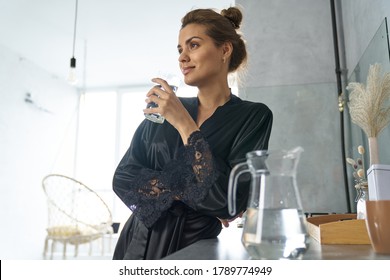 Low angle of smiling female in black dressing gown standing in kitchen with glass of water