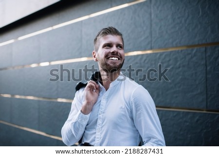 Low angle of smiling handsome bearded man holding jacket over shoulder standing beside modern building  on street and looking away