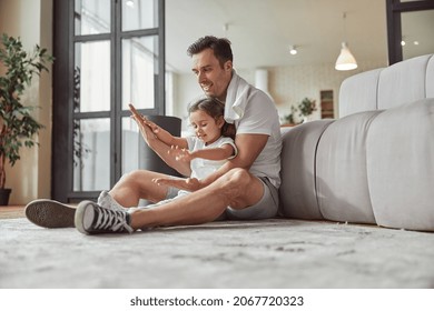 Low Angle Of Smiling Man With Little Girl Sitting On Floor In Living Room And Having Fun