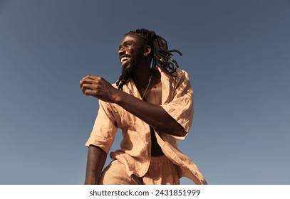 Low angle of smiling young African American male with afro braid hairstyle and in summer dress, looking away while standing alone against cloudless blue sky in sunny daylight - Powered by Shutterstock