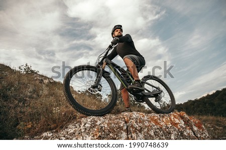 Low angle of sportive male bicyclist riding electronic mountain bike and observing environment from stony hill against cloudy sky