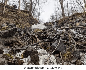 Low angle of sticks clogging up a cascading waterfall in the forest on the rocky Niagara Escarpment during the fall. A man carefully climbs down the wet hill beside the rushing stream into the valley.