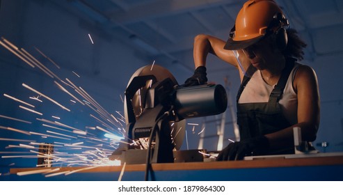 Low angle of strong female laborer in hardhat cutting metal with professional miter saw in modern factory workshop