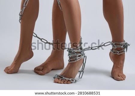 Low angle studio shot of a man and woman's bare legs and feet. Standing on a white background, their ankles are bound together by heavy metal chains, symbolizing a strong bond or restriction.