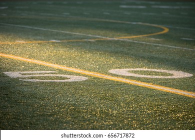 Low Angle Telephoto Perspective Of Fifty Yard Line Center Field Of Professional Or High School Football Artificial Turf Field Painted With Yellow And White Stripe In Dramatic Game Time Sunset Sunlight